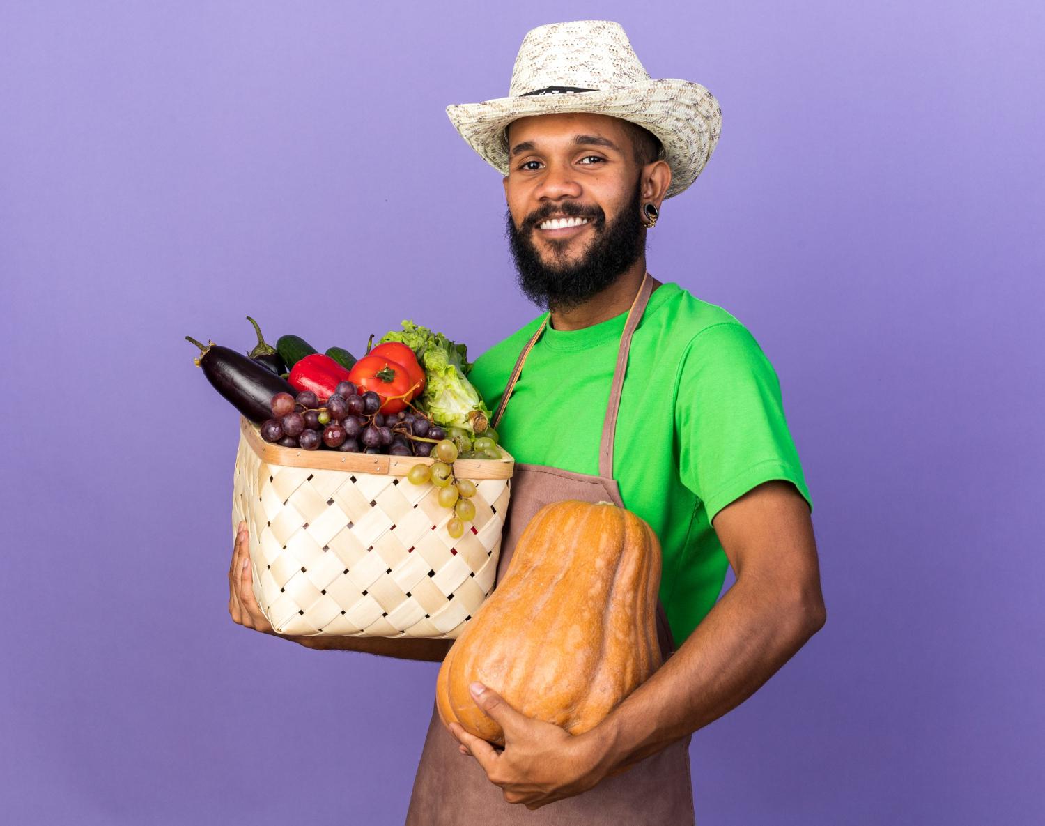smiling-young-gardener-afro-american-guy-wearing-gardening-hat-holding-vegetable-basket-with-pumpkin